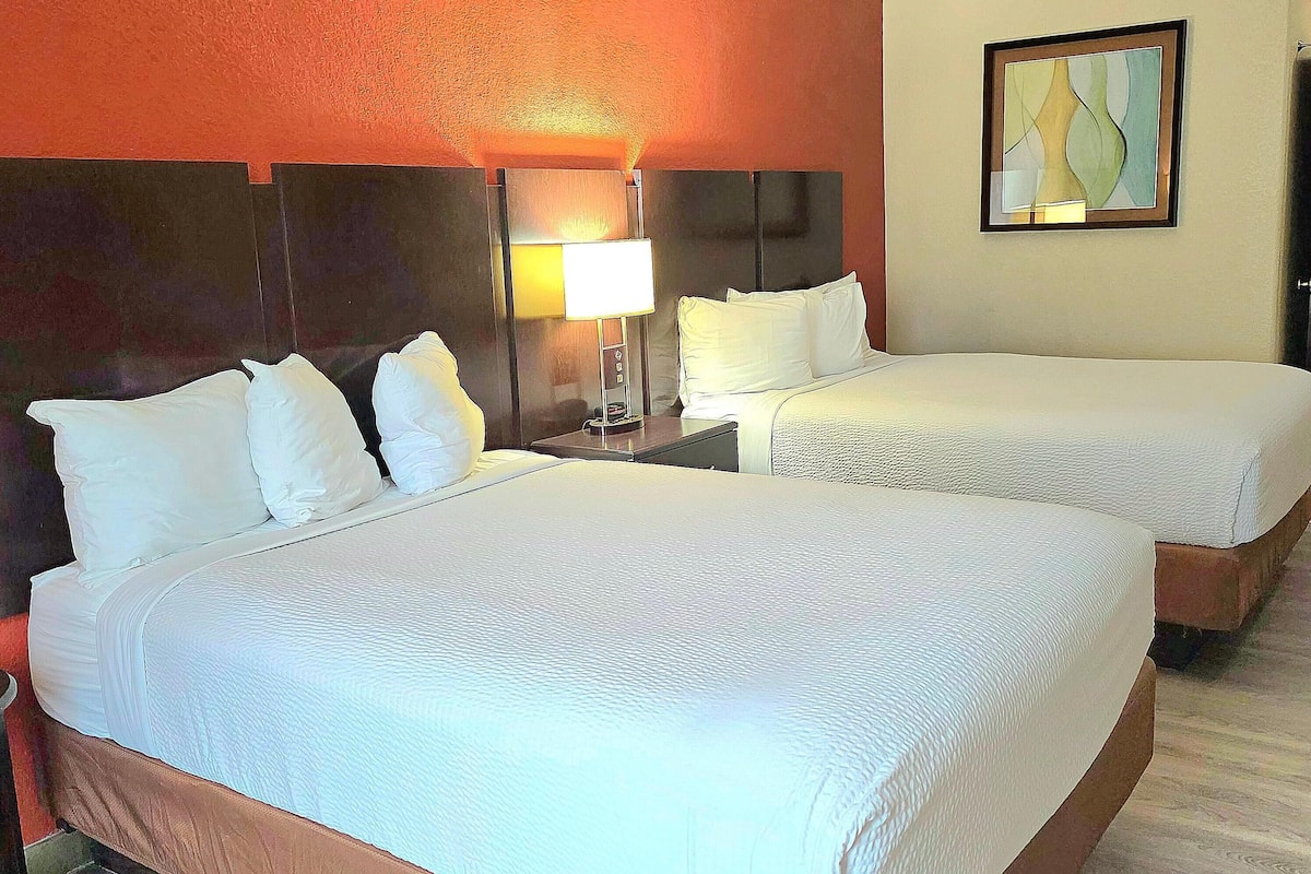 Una habitación de hotel con dos camas, una mesita de noche con lámpara y un cuadro en la pared.