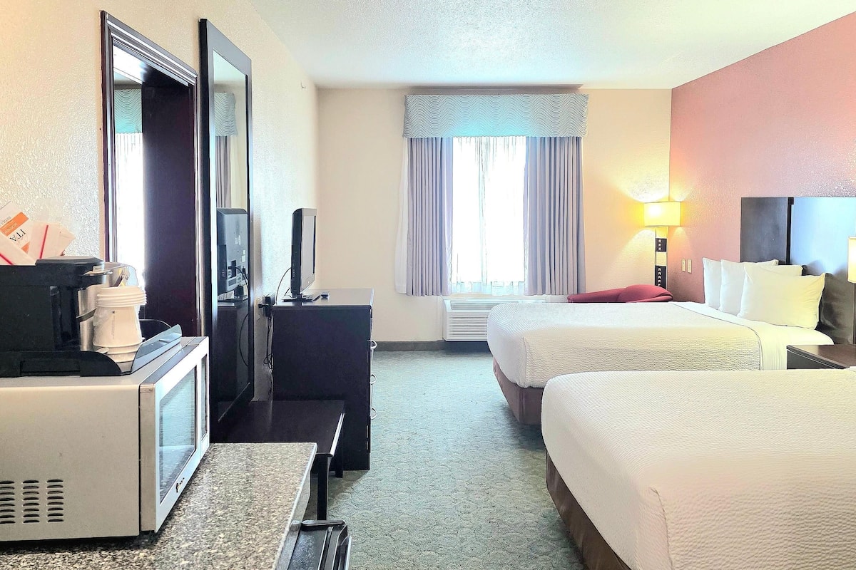 Habitación de hotel con dos camas, un escritorio con computadora, un microondas, un televisor, una ventana con cortinas y una lámpara de pared.