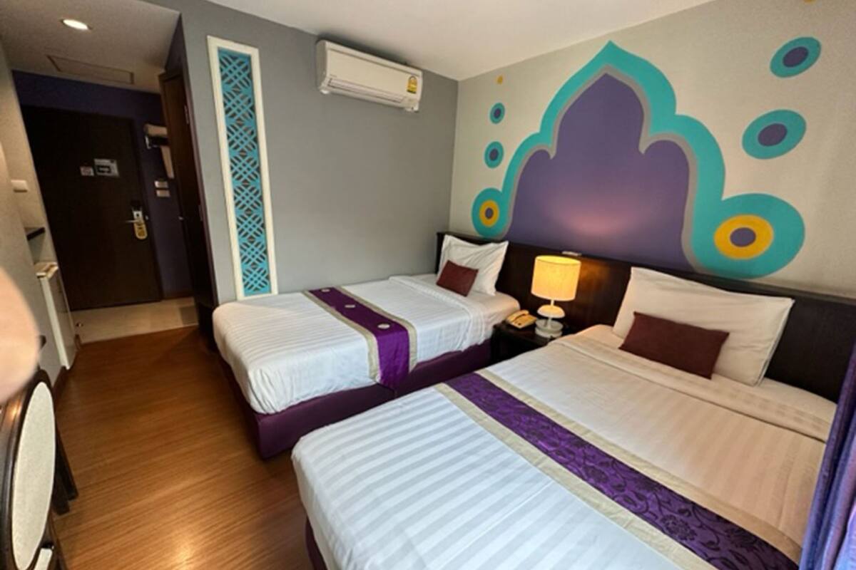 A hotel room with two beds, a nightstand with a lamp, an air conditioning unit, and a decorative wall with a geometric pattern.