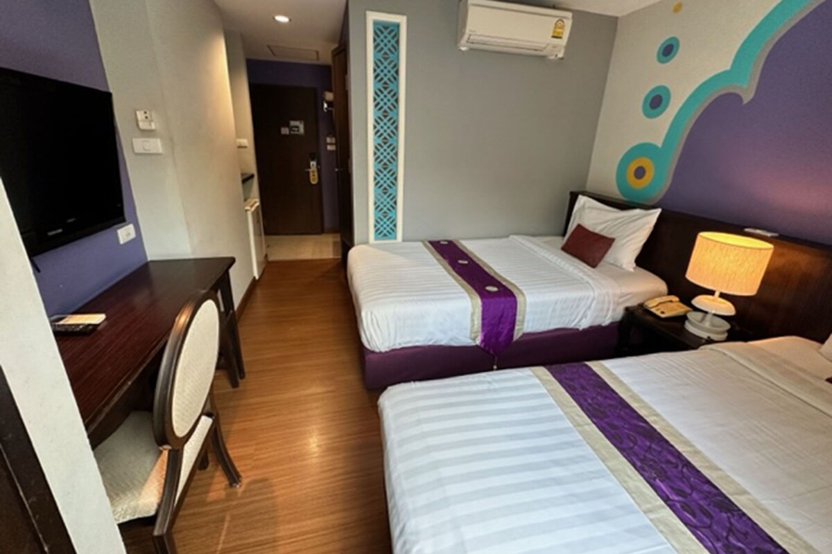 A hotel room with two beds, a desk with a chair, a TV, and an air conditioning unit.