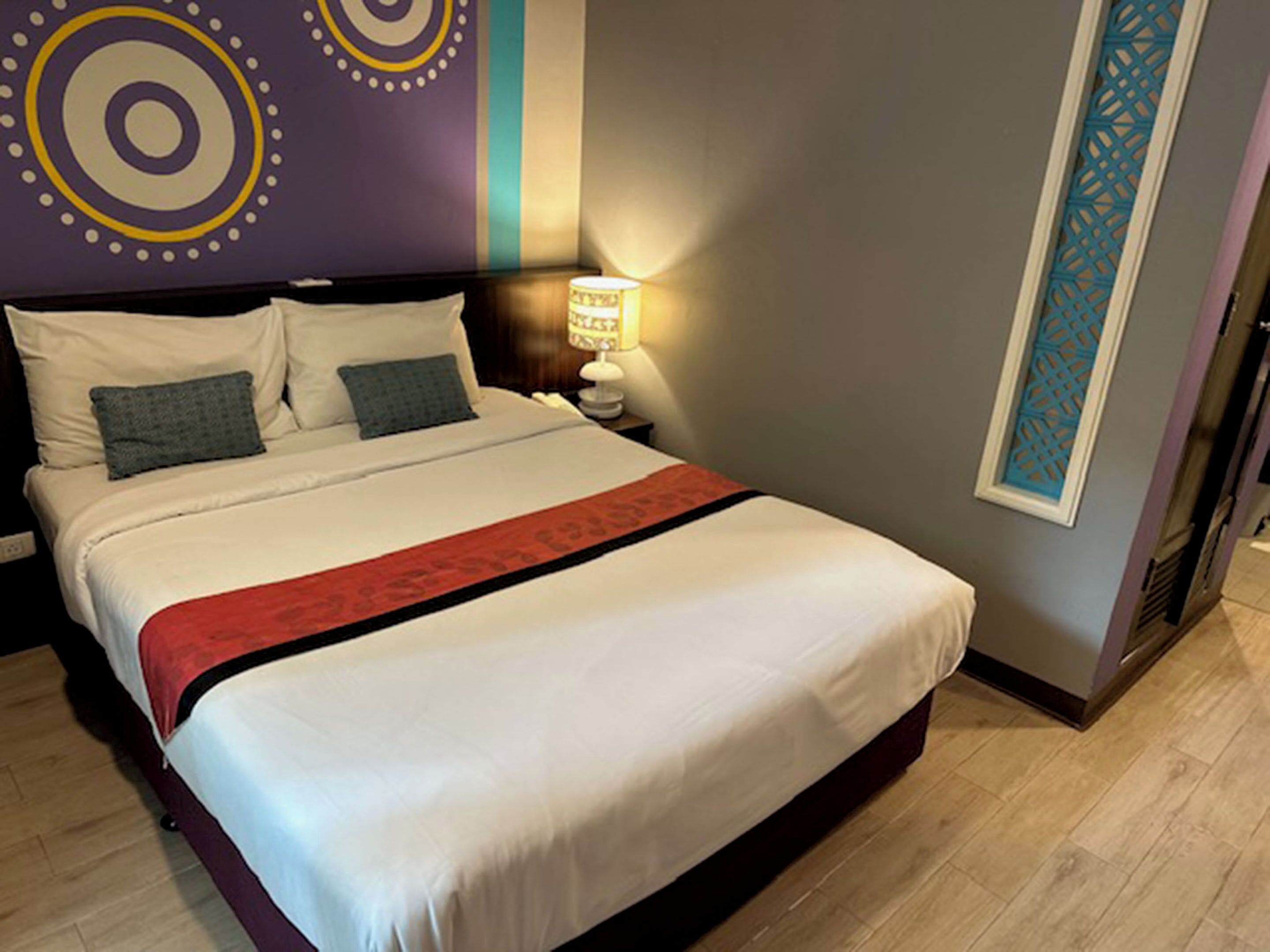 A neatly made bed with white linens and a red accent, a bedside lamp, and a decorative wall with circular patterns.