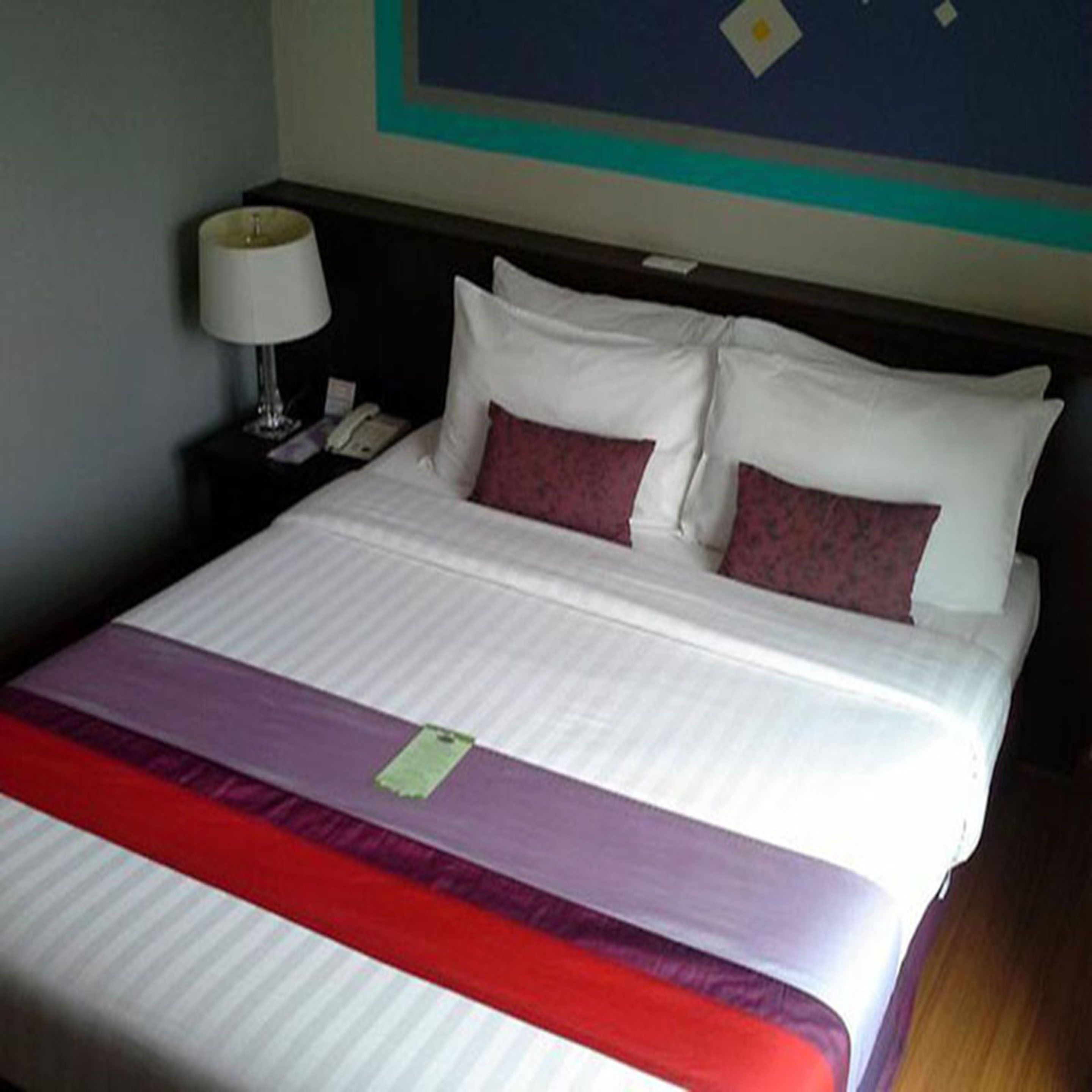 A neatly made bed with white and red bedding, a nightstand with a lamp and phone, and a wall with a geometric design.
