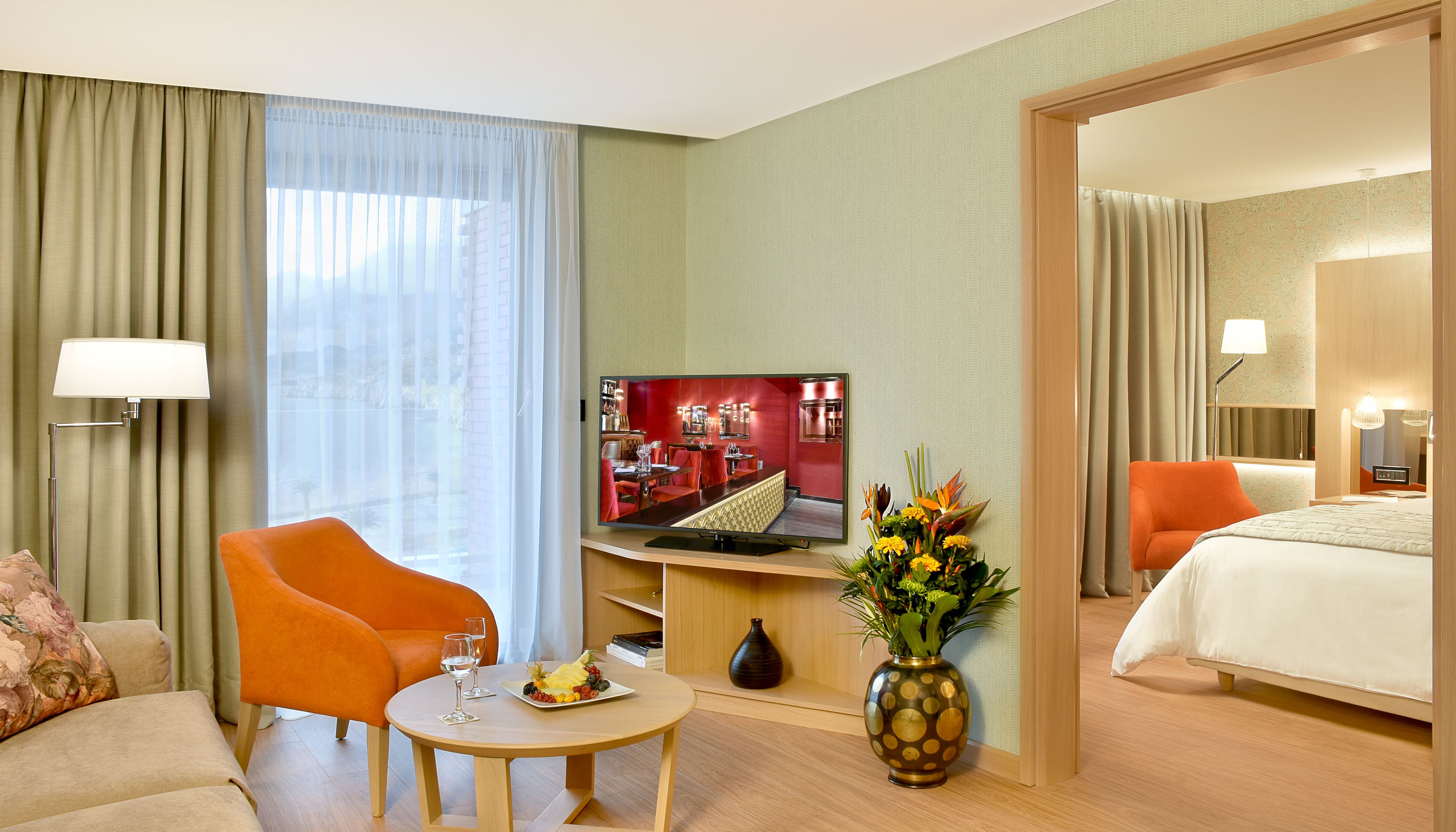 A hotel room with a bed, orange armchair, round table with a plate of fruit and glasses, a flat-screen TV, and a vase with flowers.