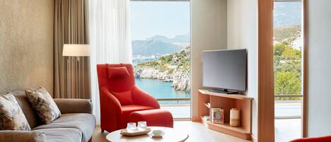A modern hotel room with a sofa, a red armchair, a television, and a view of the sea.