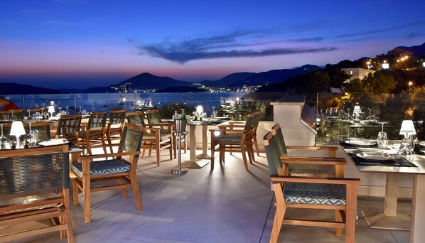 An outdoor dining area with tables and chairs, overlooking a body of water and mountains at dusk.
