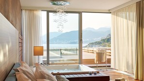 Habitación de hotel moderna con un ventanal grande, suelo de madera, sofá, una mesita y vistas al mar y a la montaña.