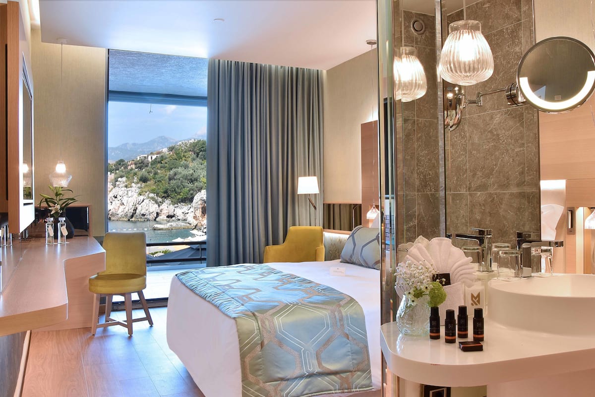 Una habitació d'hotel moderna amb un llit gran, una zona de seients amb una cadira groga, un lavabo de bany rodó i vistes al mar.