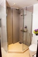 Un box de ducha con paredes de vidrio, un cabezal de ducha manual, una puerta de vidrio y un pequeño estante con artículos de tocador.