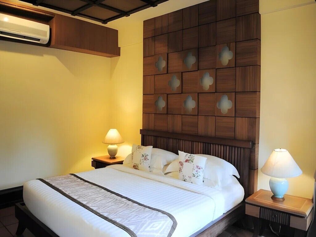 A hotel room with a bed, wooden headboard, two bedside tables with lamps, and an air conditioning unit.
