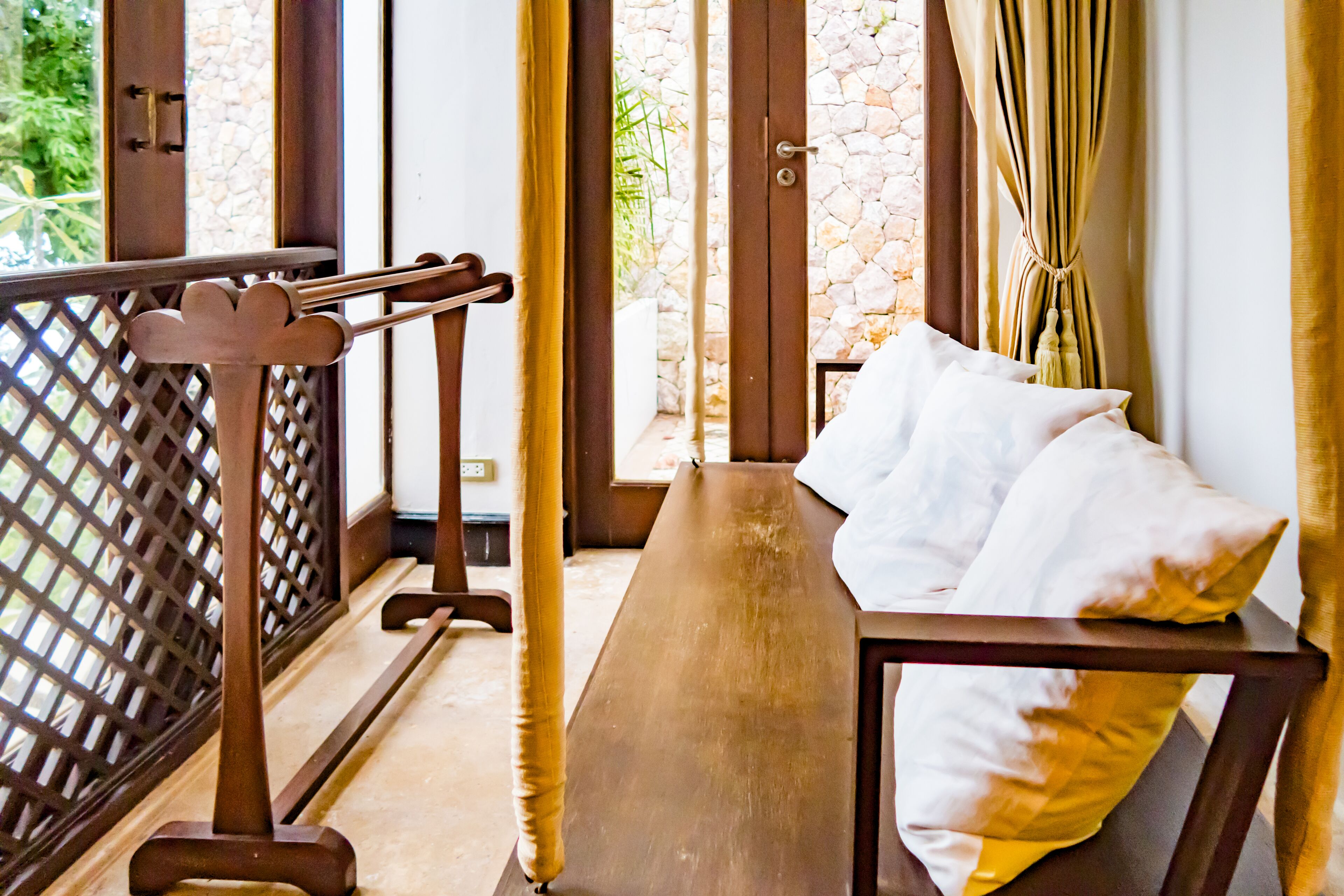 A room with a wooden bench, a bed with white pillows, and a view of greenery through a window with curtains.