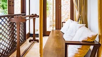 A room with a wooden bench, a bed with white pillows, and a view of greenery through a window with curtains.