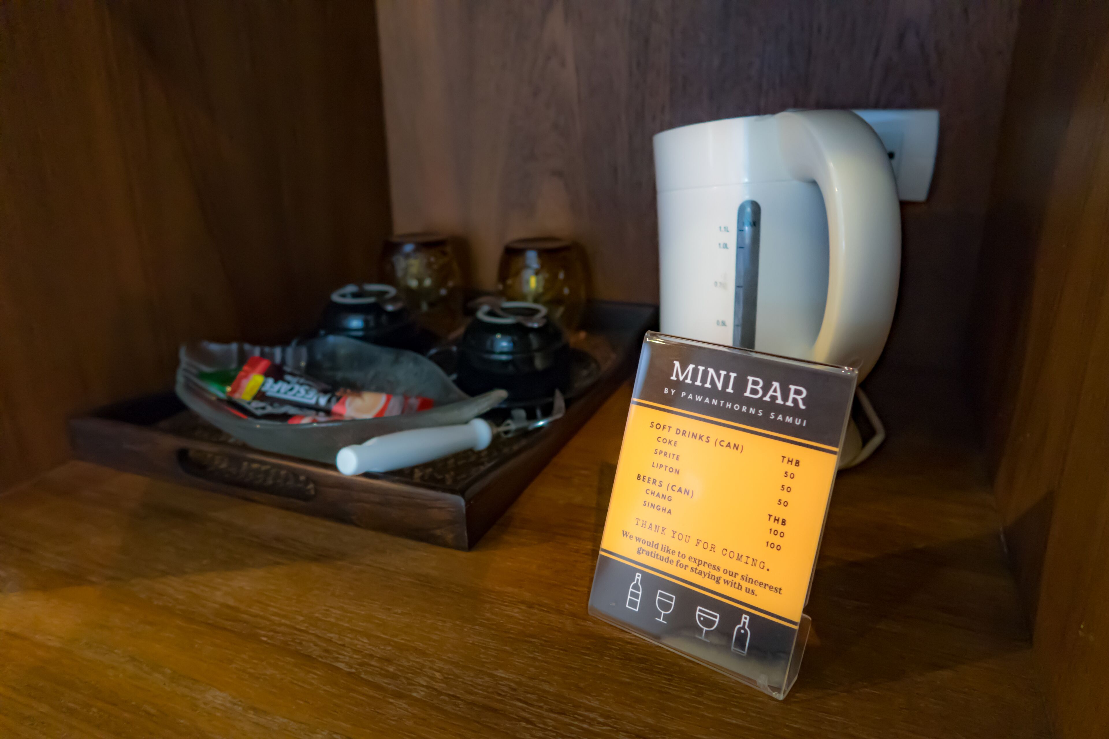 A mini bar with a sign listing soft drinks, Coke, Sprite, Lipton, beers, and Singha with their prices.