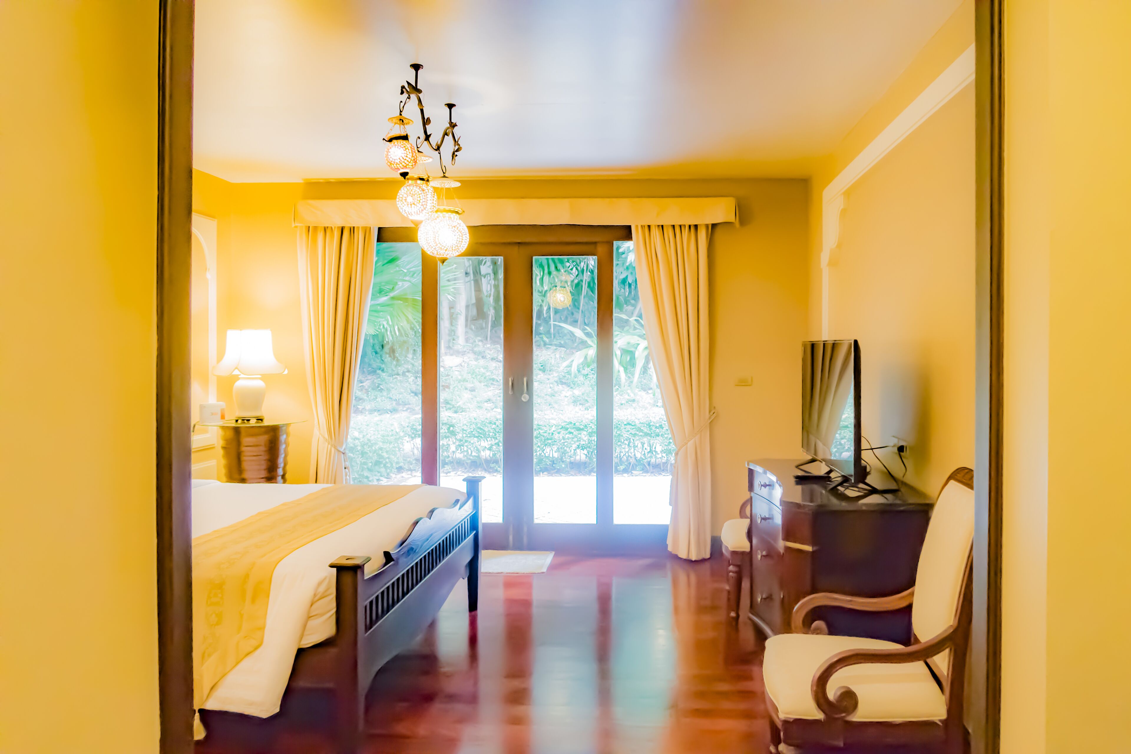 A hotel room with a bed, a desk, a chair, a lamp, and a view of greenery outside.