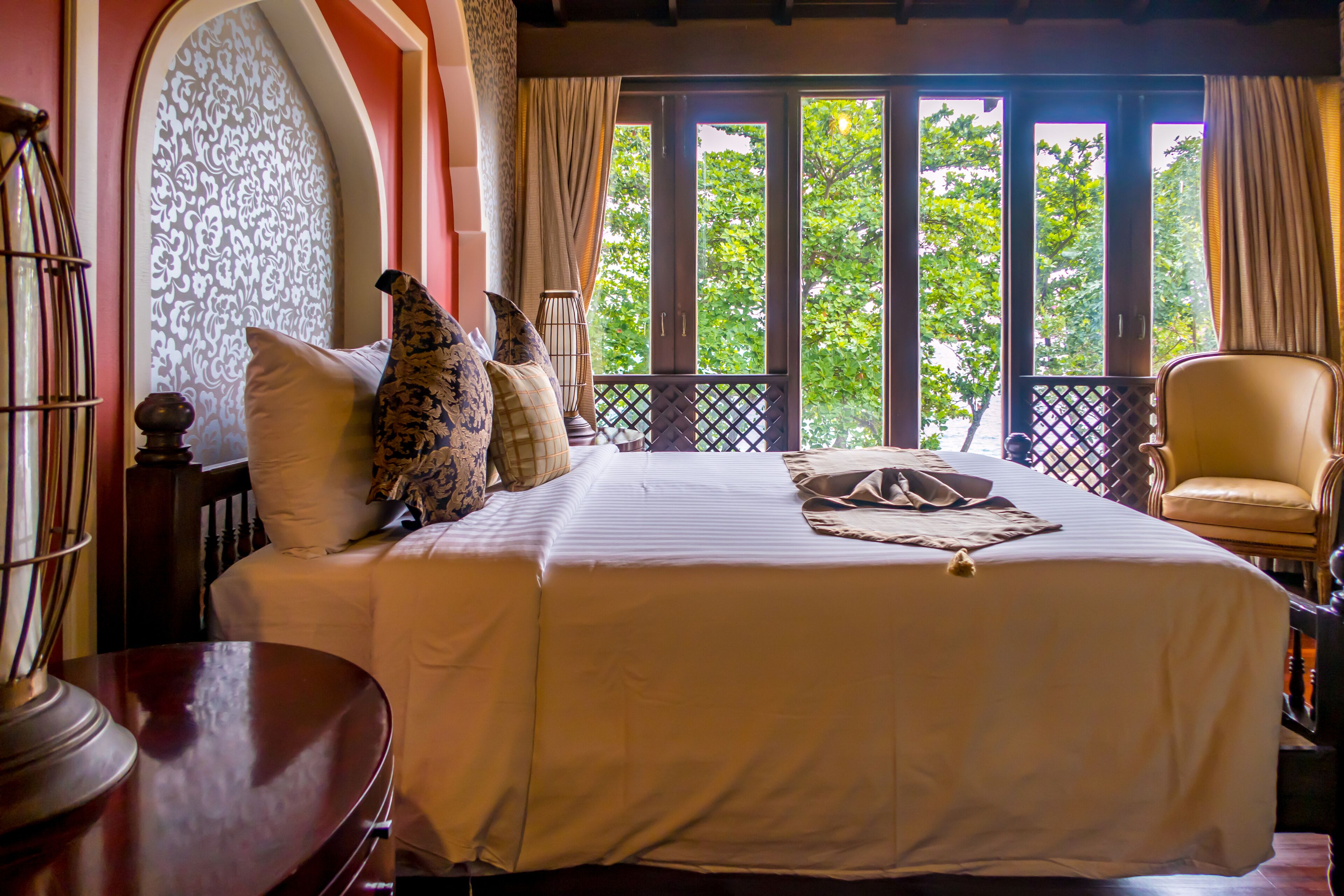 A hotel room with a large bed, a wooden chair, and a view of trees through the window.