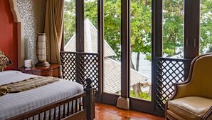 Quarto com cama, uma cadeira, mesa de cabeceira, abajur e vista do exterior através de grandes janelas.