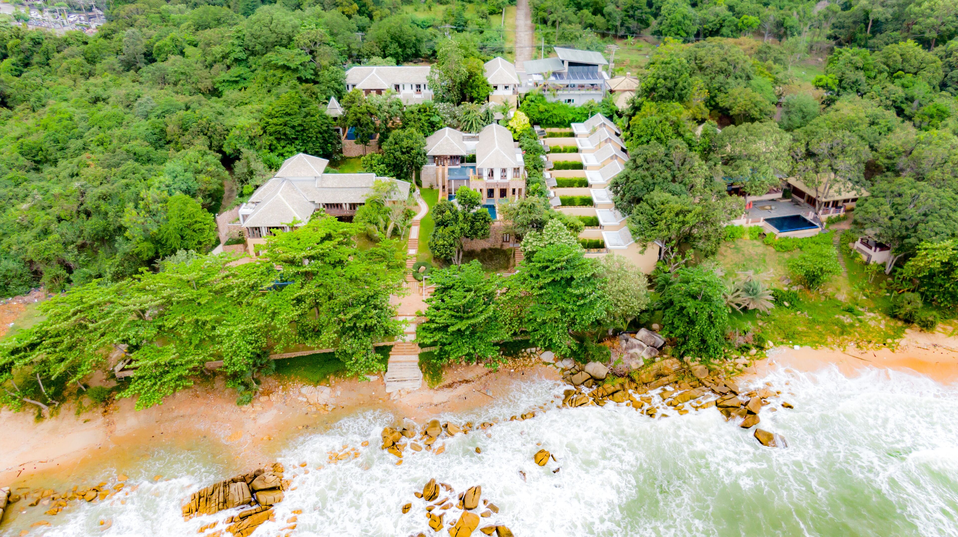An aerial view of a coastal property with multiple houses, a beach, and a rocky shoreline.