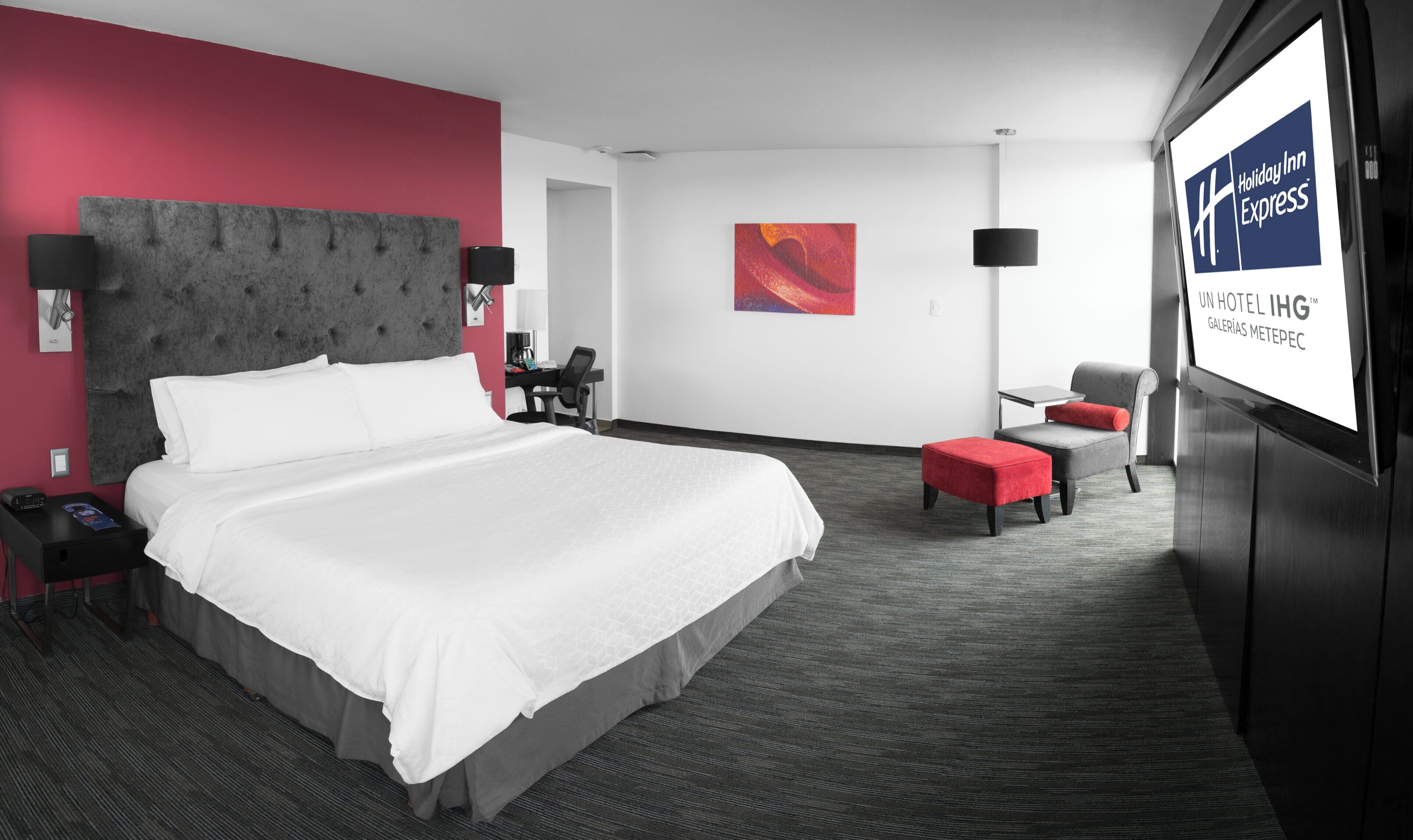 Ett hotellrum med en stor säng, ett skrivbord, en stol och en tv-skärm som visar hotellets namn.
