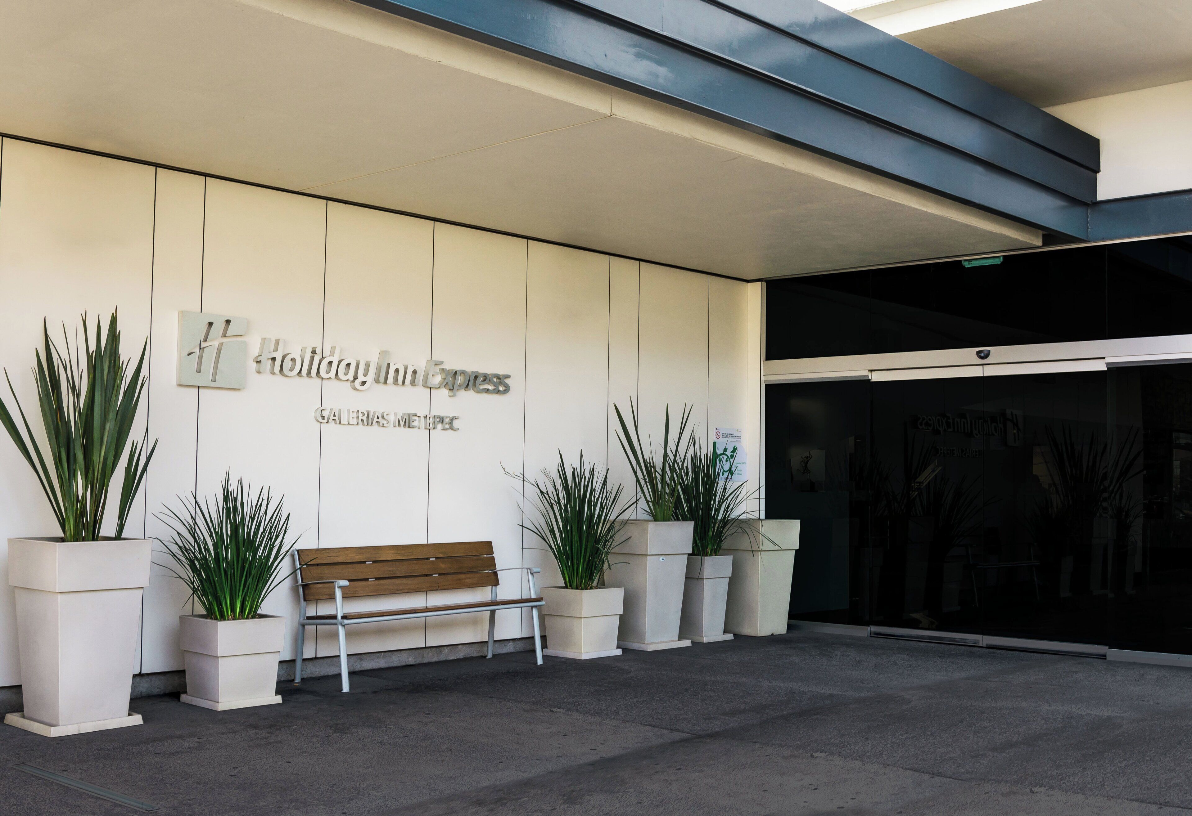 Entrada de hotel con un banco, plantas en macetas y un cartel que dice 'Holiday Inn Express'.
