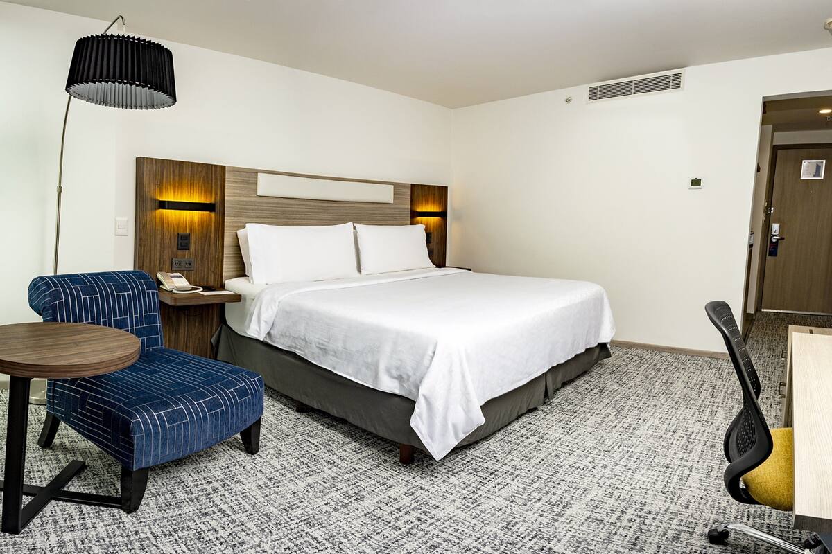 A hotel room with a bed, a desk, a chair, a lamp, and a small table.