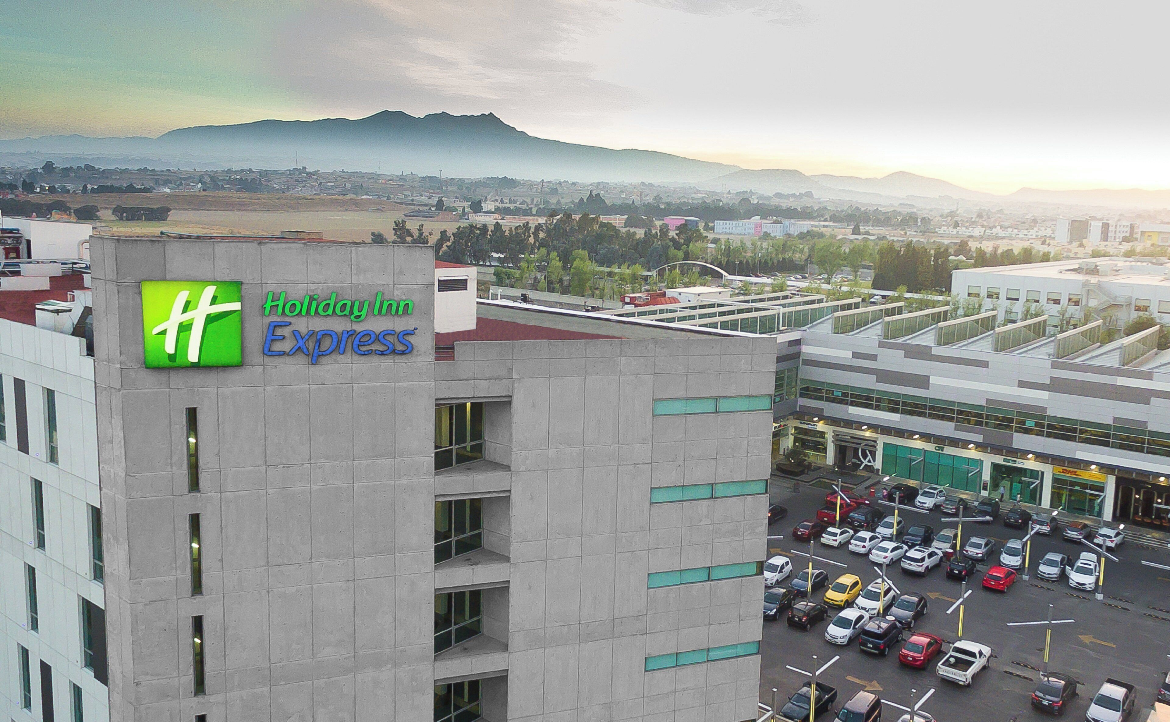 Edificio de Holiday Inn Express con vista a un estacionamiento y montañas a la distancia.