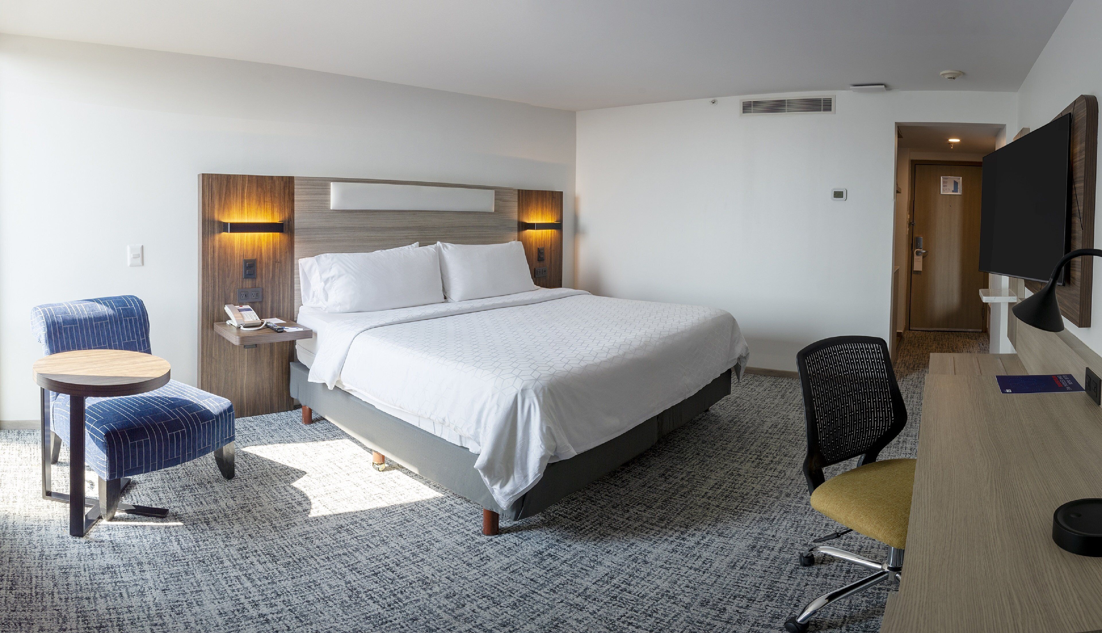 A hotel room with a bed, a desk, a chair, a TV, and a small table with a phone.