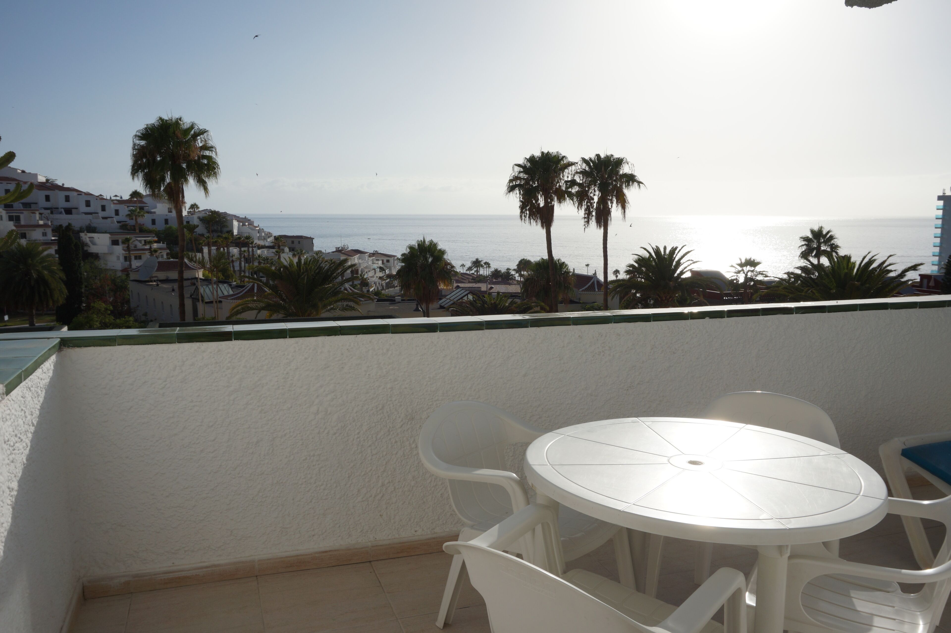 Un balcón con una mesa y sillas blancas, con vistas a un pueblo costero con palmeras y edificios.