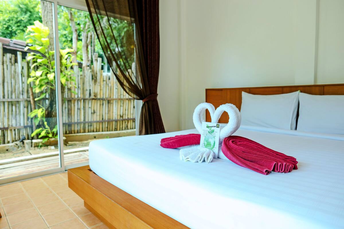 A hotel room with a bed, a wooden headboard, and a view of a bamboo fence and greenery outside.
