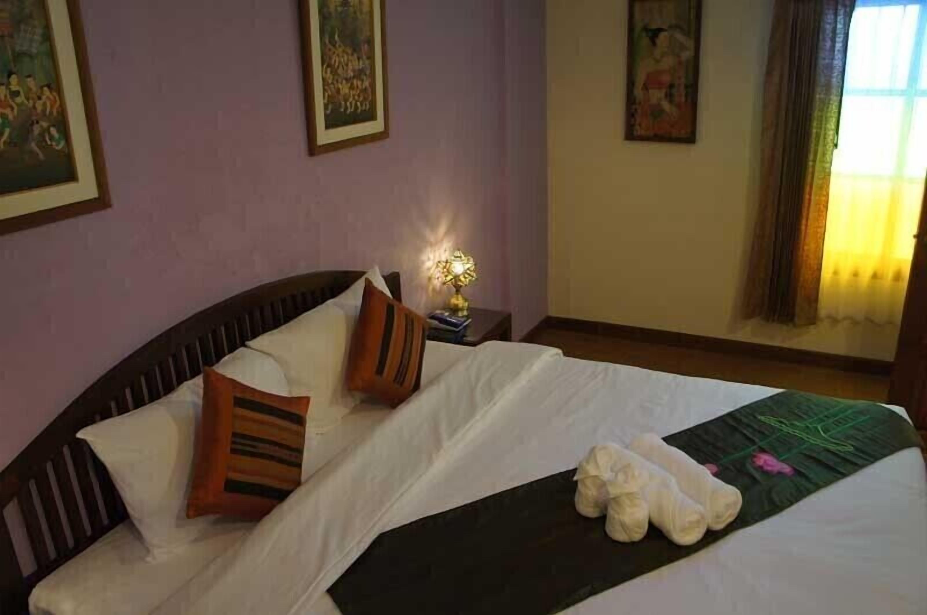 A neatly made bed with pillows, a bedside lamp, and rolled towels on a bed in a room with purple walls and artwork.