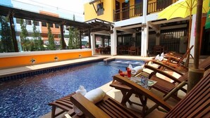 Un área de piscina con sillones de madera y un edificio amarillo al fondo.