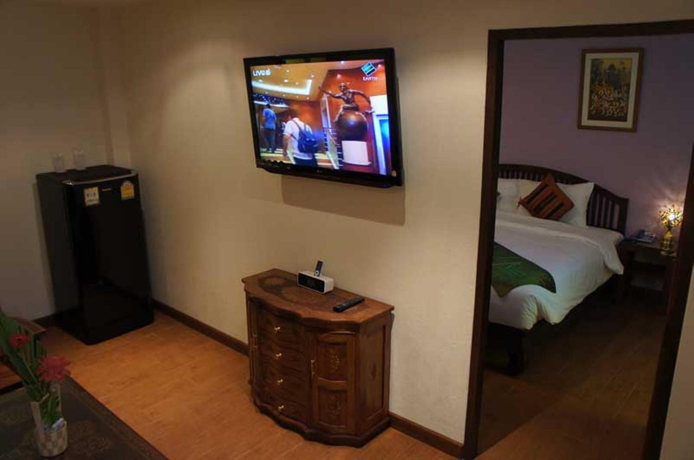A hotel room with a television mounted on the wall, a wooden cabinet, a bed with pillows, and a nightstand with a lamp.