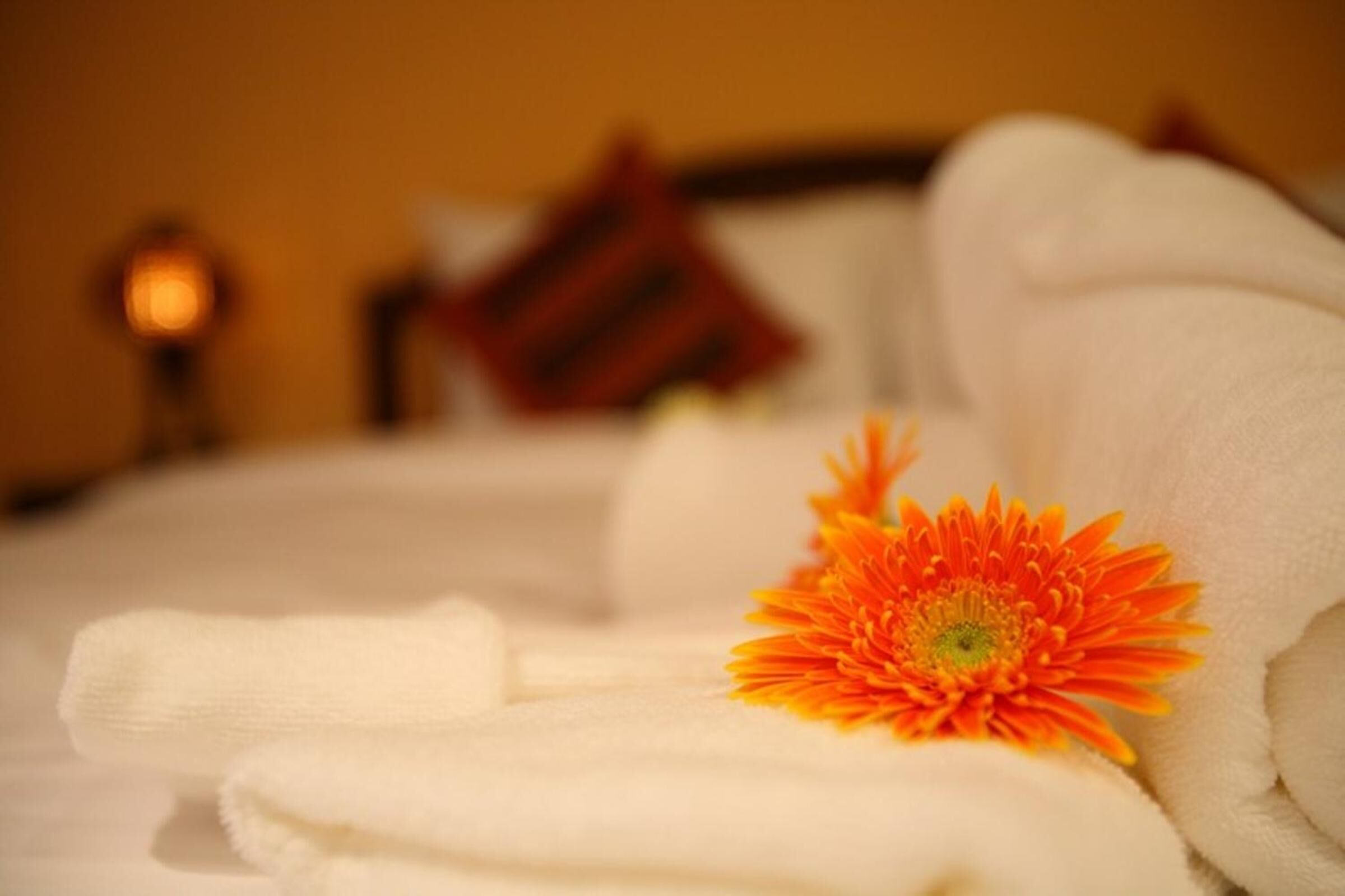 A neatly folded white towel with a single orange flower on it, placed on a bed with a red pillow in the background.