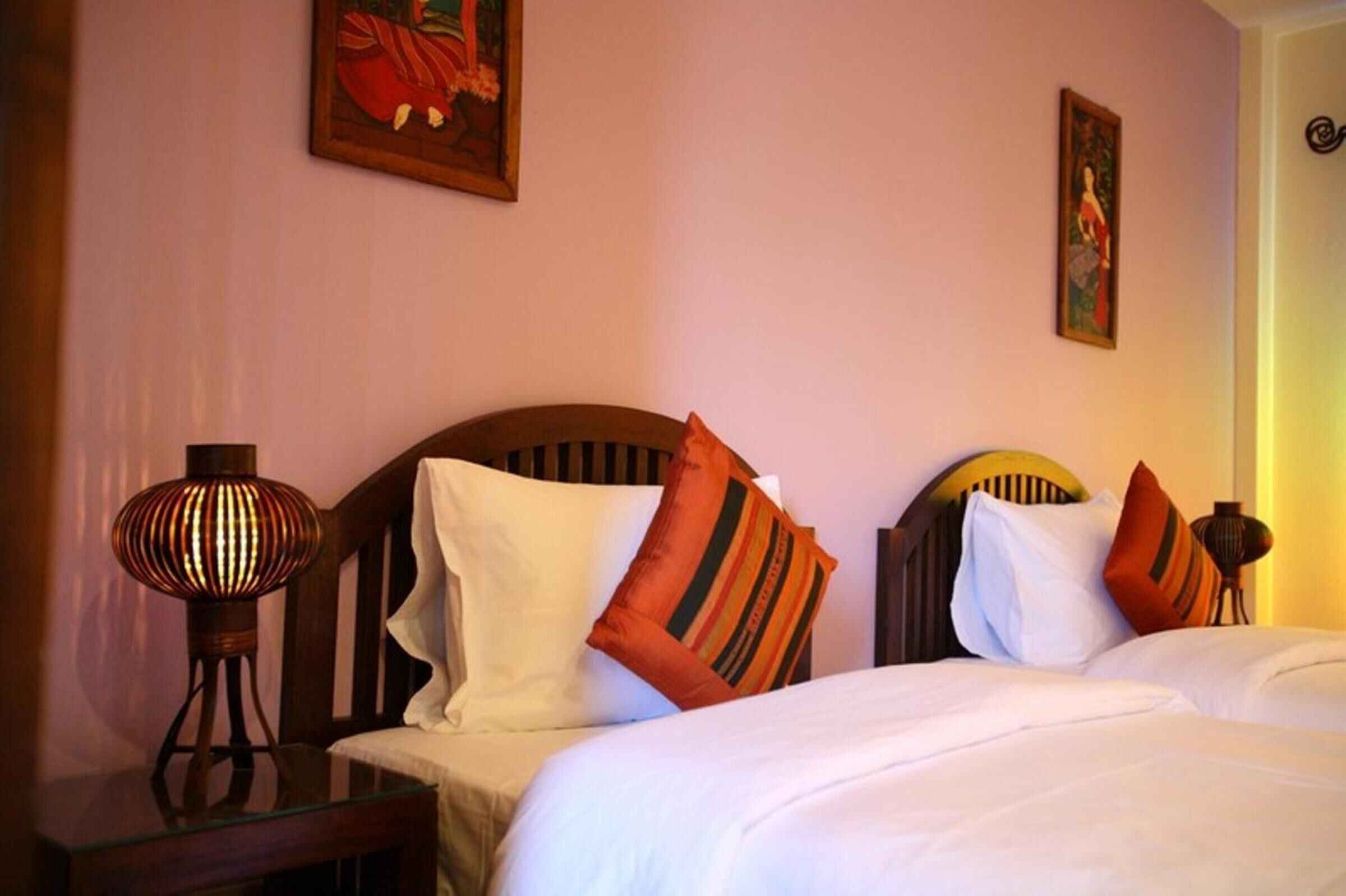A hotel room with two beds, each with a wooden headboard, white bedding, and striped pillows. There is a table lamp on the left side of the room.