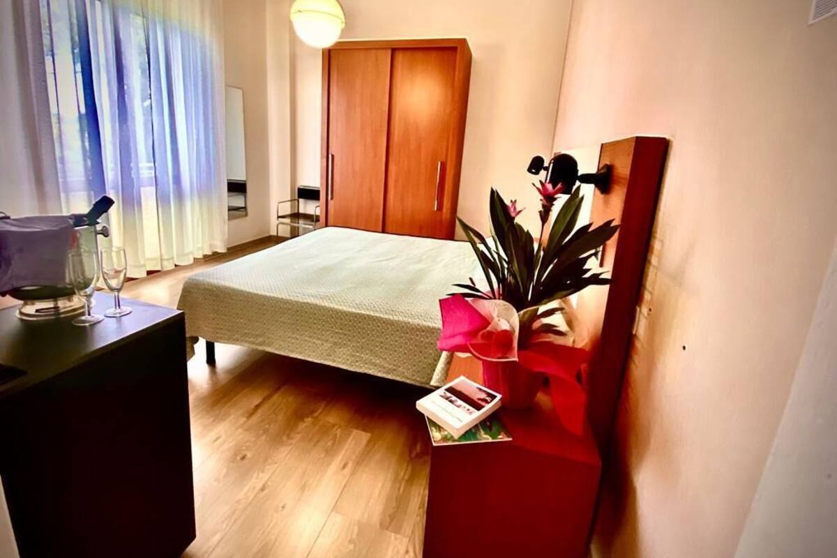 A hotel room with a bed, a wooden wardrobe, a bedside table with a lamp, a potted plant, and a window with sheer curtains.
