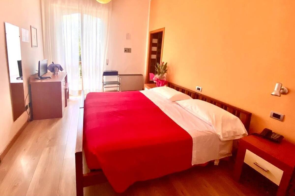 A hotel room with a bed, a desk, a chair, a small table, and a potted plant.
