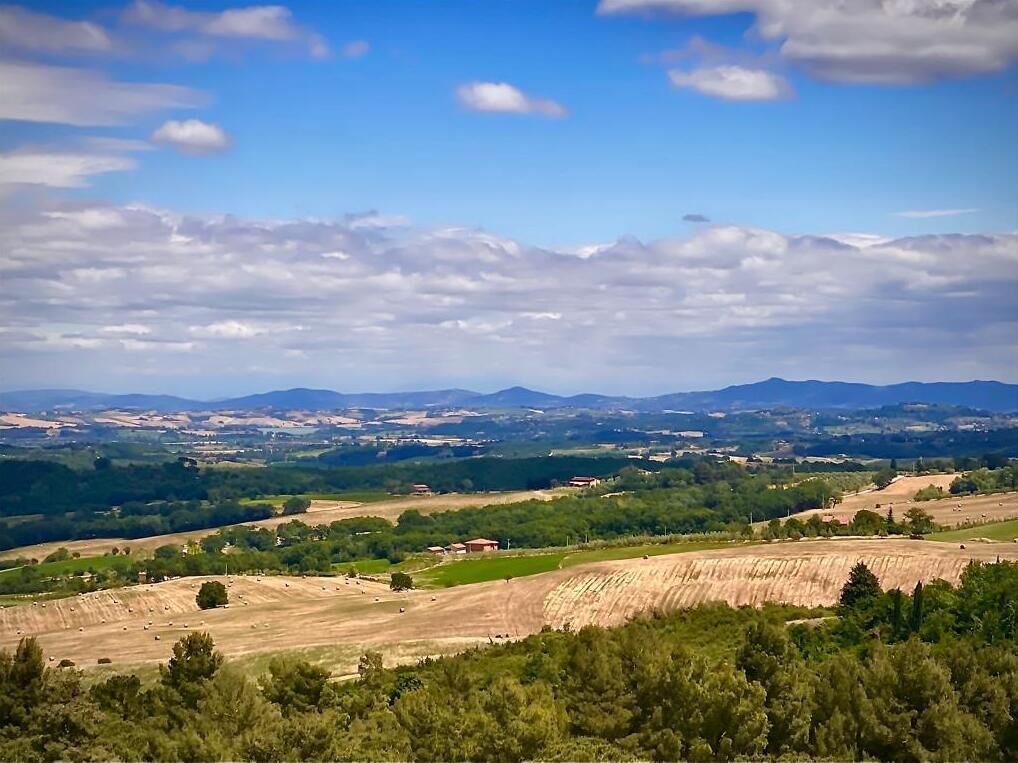 Een panoramisch uitzicht op een landelijk landschap met glooiende heuvels, velden en verre bergen, onder een gedeeltelijk bewolkte lucht.