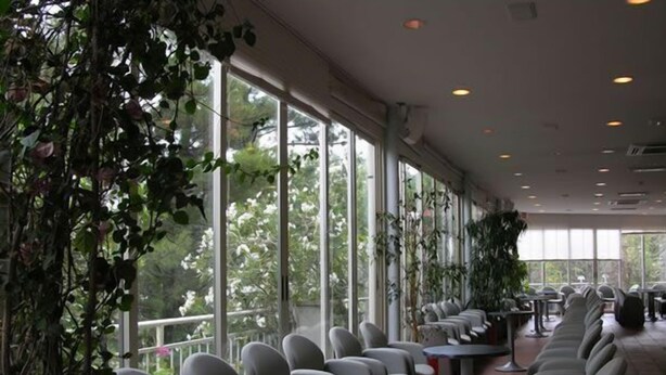 Um espaço amplo e interno com fileiras de cadeiras e mesas, janelas grandes e plantas.