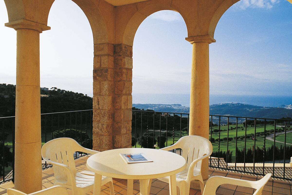 A terrace with a table and chairs, overlooking a scenic landscape with hills and a body of water.