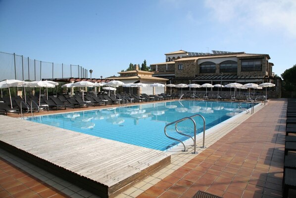 A swimming pool with sun loungers and umbrellas, surrounded by a wooden deck and a stone building in the background. - Hapimag Resort Mas Nou (Castell-Platja d'Aro)