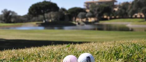 A golf course with two golf balls on the grass, a pond, and buildings in the background.