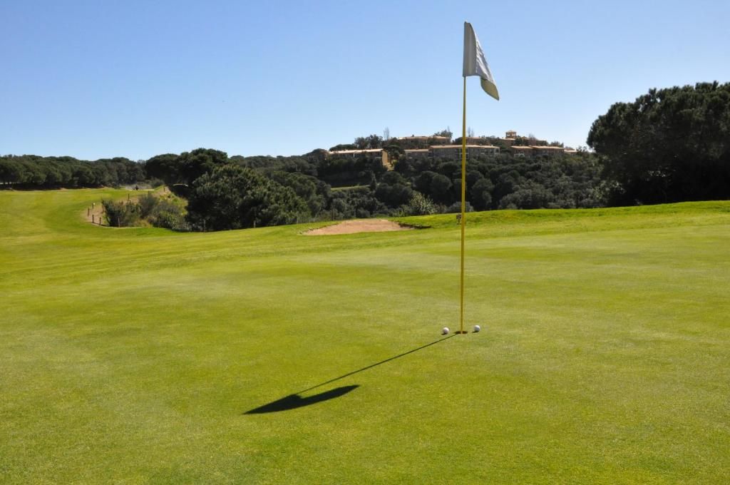 Campo de golfe com haste de sinalização, céu limpo e colina com edifícios ao fundo.