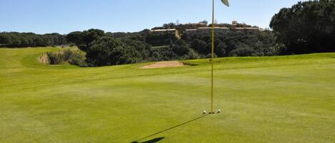 Campo de golfe com haste de sinalização, céu limpo e colina com edifícios ao fundo.