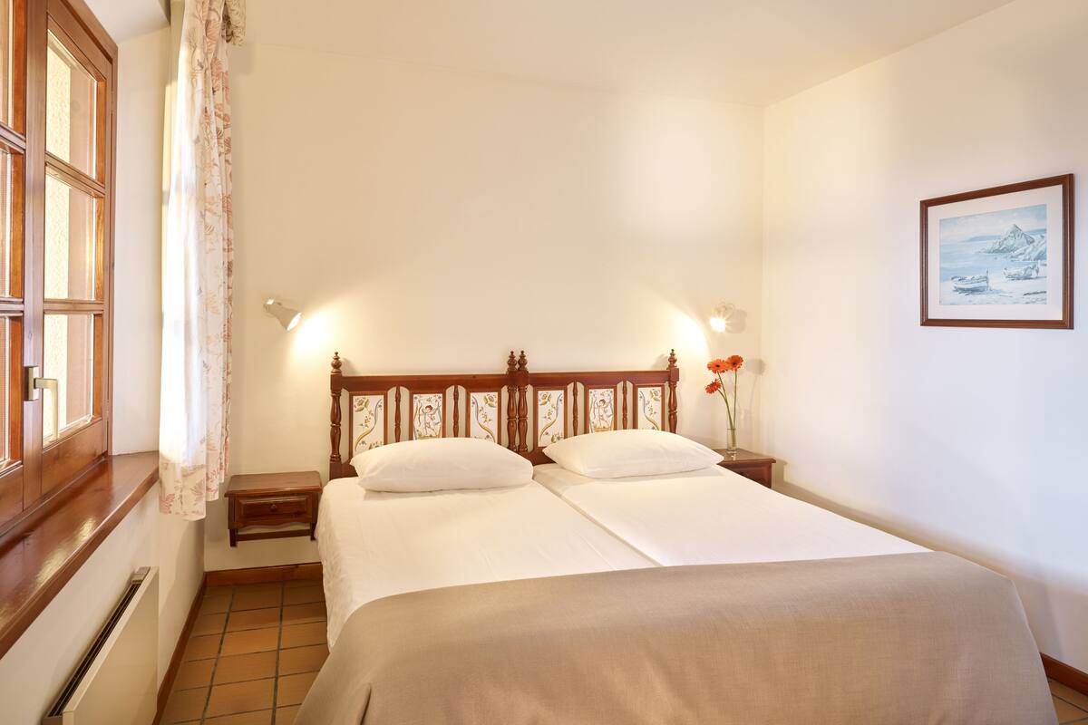 A bedroom with two beds, a wooden headboard, a nightstand with a vase of flowers, a framed picture on the wall, and a window with curtains.