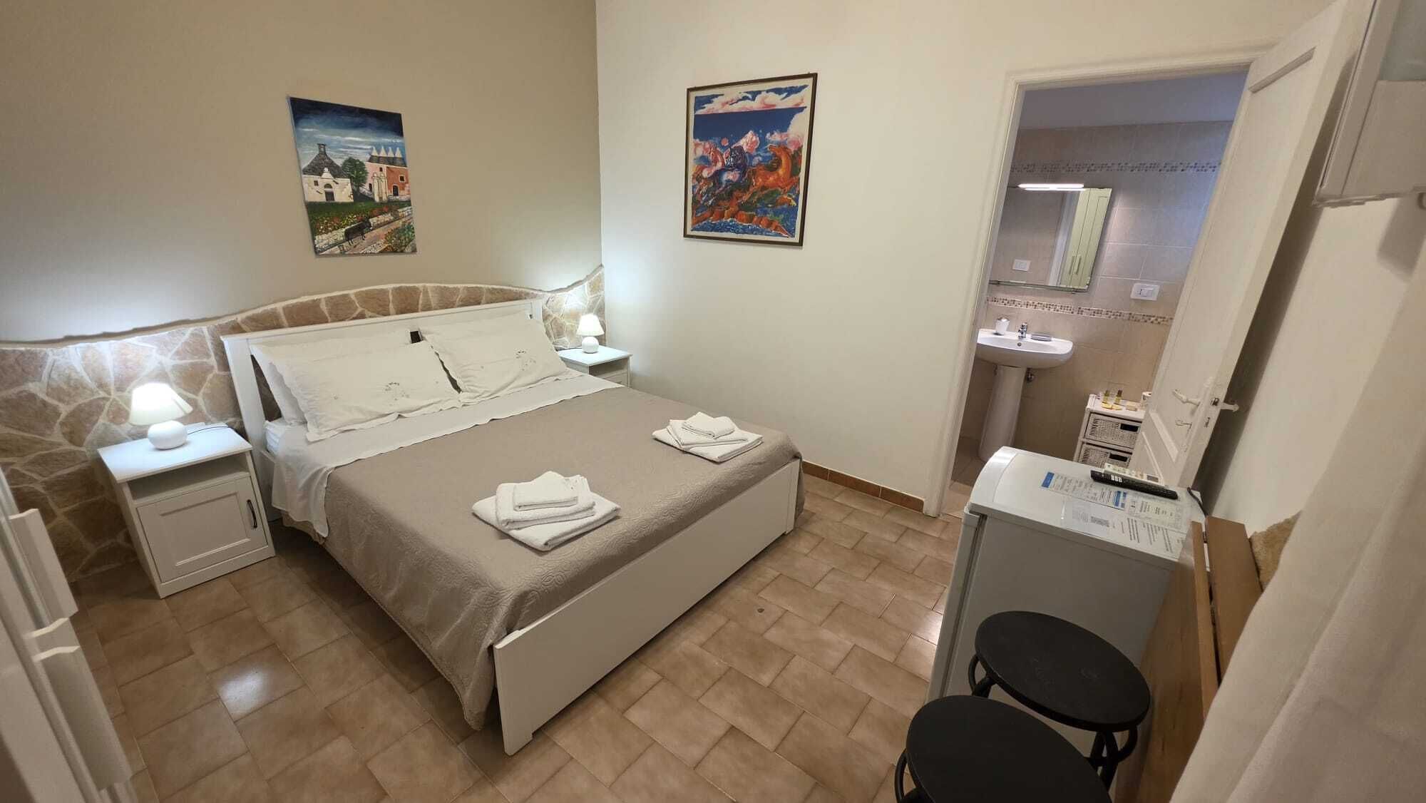 A bedroom with a bed, bedside tables, a nightstand, a desk, a chair, a mirror, and a bathroom with a sink and shower.