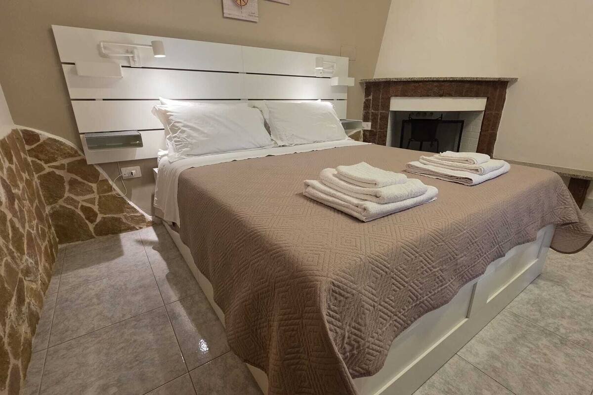 A bedroom with a bed, a nightstand, a fireplace, and a stone wall.