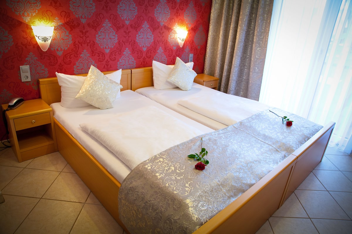 A double bed with white linens, two decorative pillows, a bedside table with a phone, and a wall-mounted light fixture.