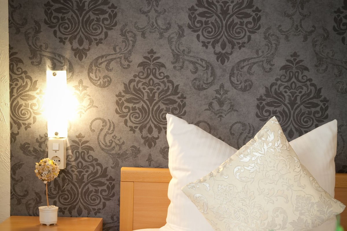 A neatly made bed with white linens and a decorative pillow, a bedside table with a lamp and a vase of flowers, and a patterned wallpaper in the background.