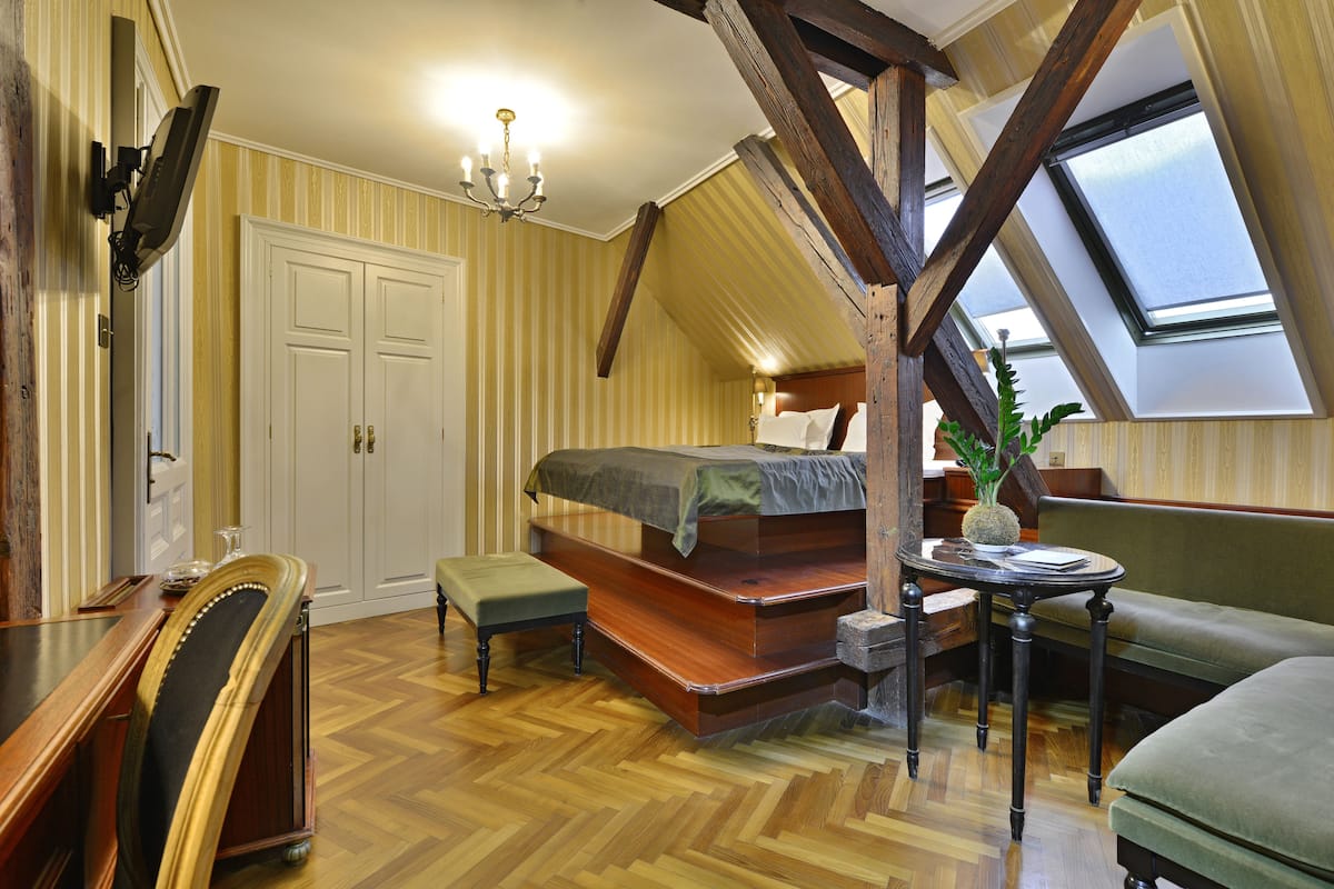 A bedroom with a loft bed, wooden beams, a chandelier, a small table, and a window.