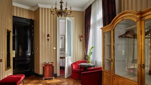 A room with a wooden herringbone floor, a red upholstered armchair, a small round table with a vase, a large wooden cabinet with glass doors, and a black door. - Gerlóczy Boutique Hotel (Budapest)