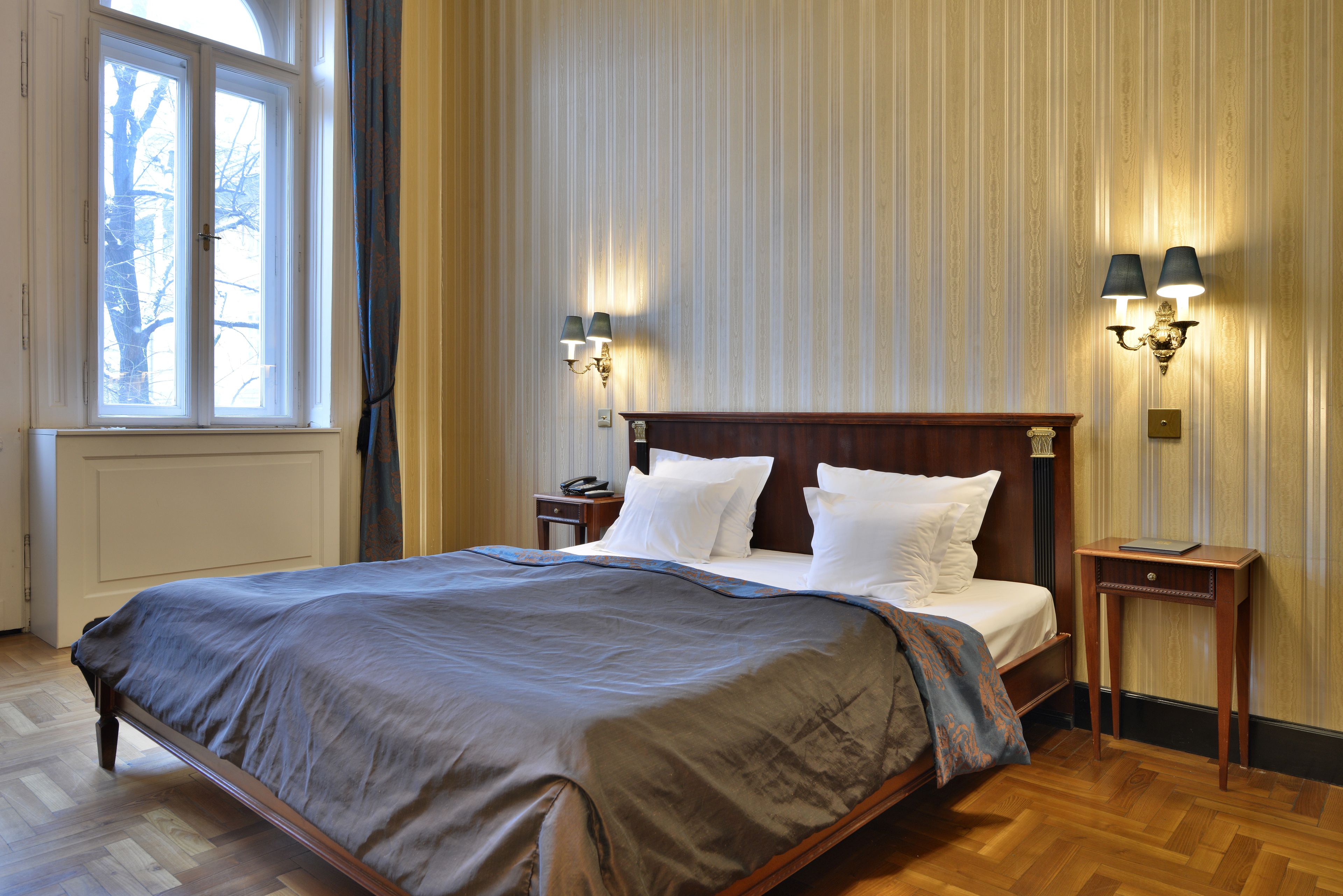 Una cama bien hecha con tablero de madera, dos mesitas de noche con lámparas, una ventana con cortinas y un radiador.