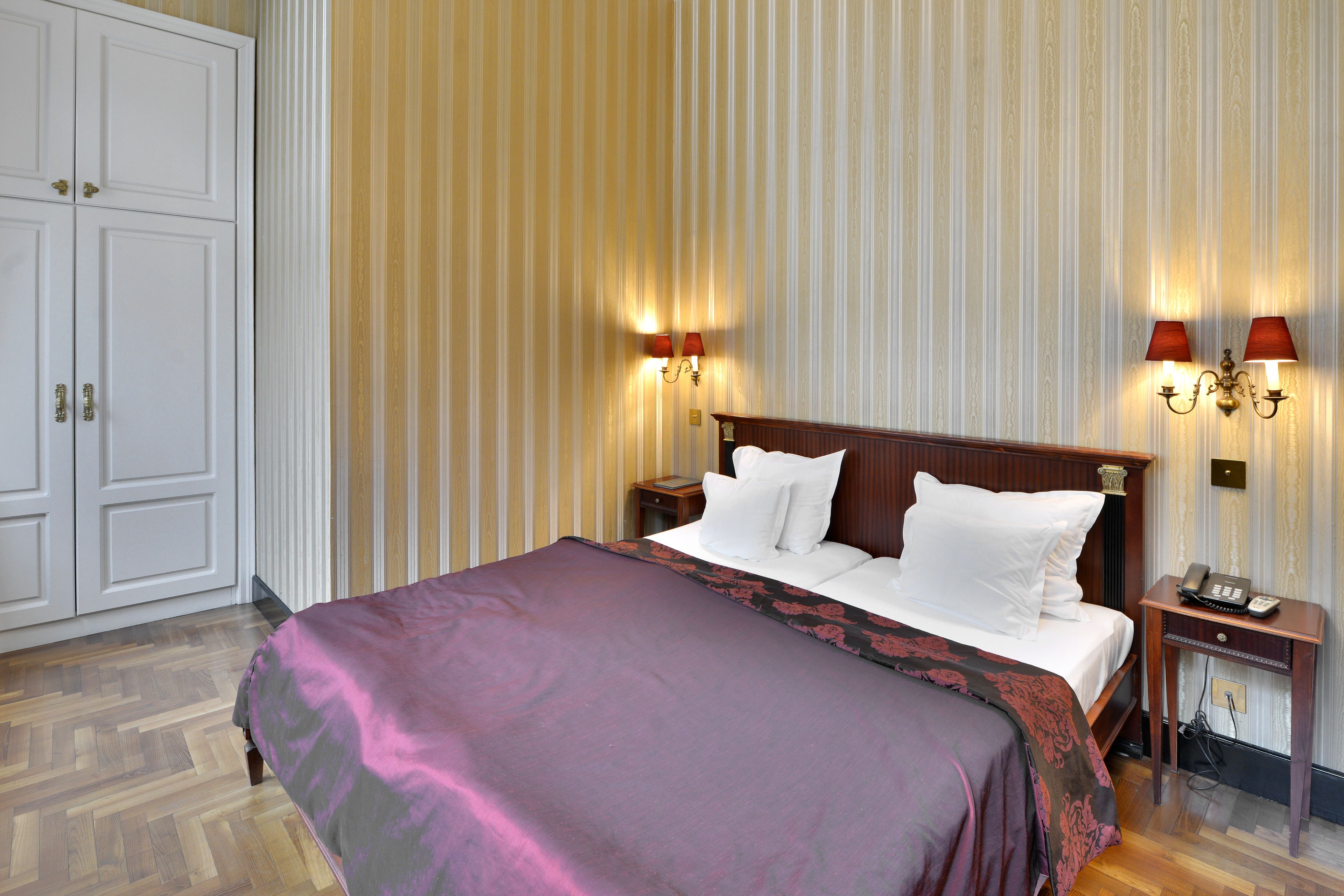 A hotel room with a large bed, a nightstand with a telephone, and two wall-mounted lamps.
